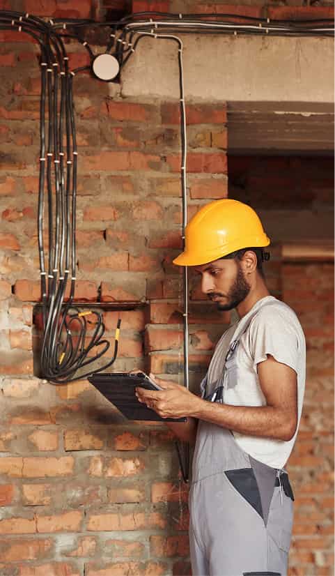 Electrician wearing a yellow hard hat inspecting electrical wiring on an unfinished brick wall using a digital tablet