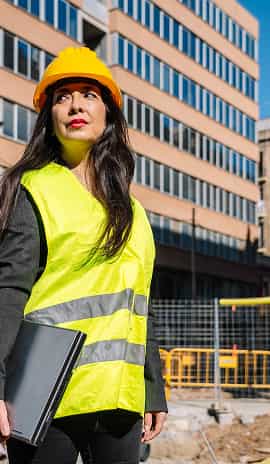 Female construction professional in a hard hat and high-visibility vest holding a laptop at an urban building site