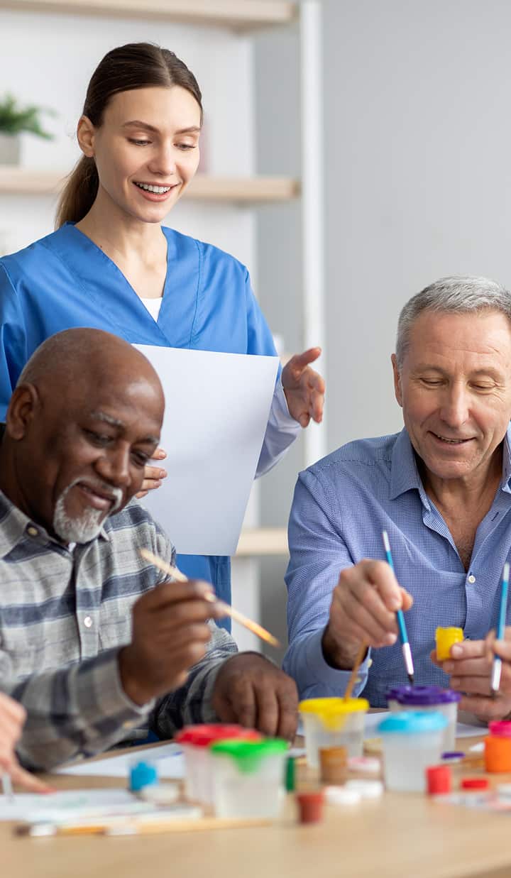 Two people are seated at a table painting with brushes and colorful paint containers, while a person in blue scrubs stands behind them holding a sheet of paper in a bright, organised room.