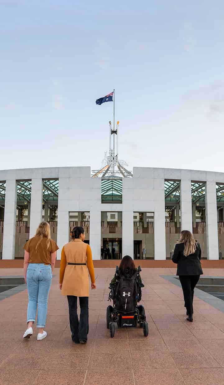 A group of four people, including one using a motorised wheelchair, walk toward the front entrance of Australia’s Parliament House in Canberra, with the Australian flag flying above the building.