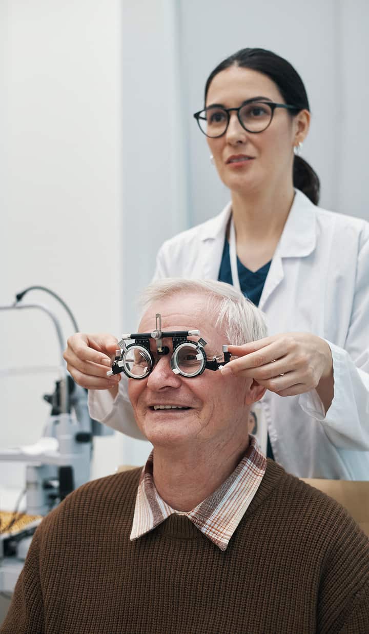 A healthcare professional in a white coat is adjusting a pair of eyeglasses on a seated patient in a clinical setting.