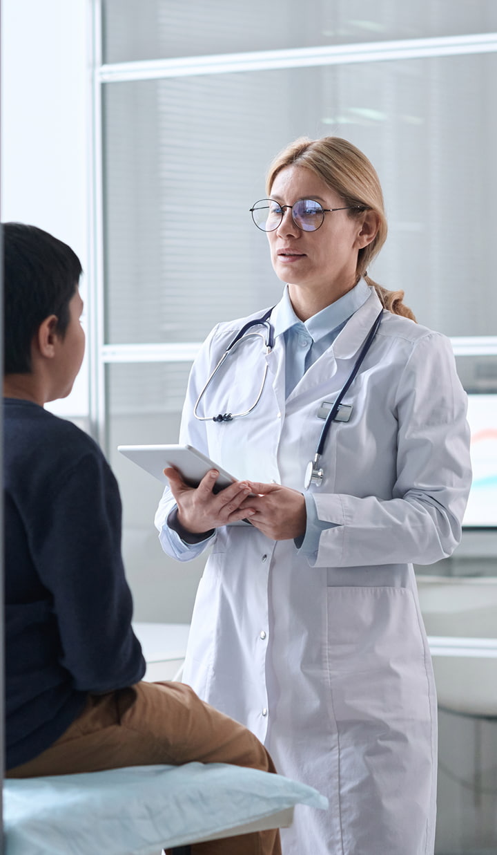 A doctor in a white lab coat with a stethoscope is holding a tablet and speaking to a seated patient in a medical examination room.