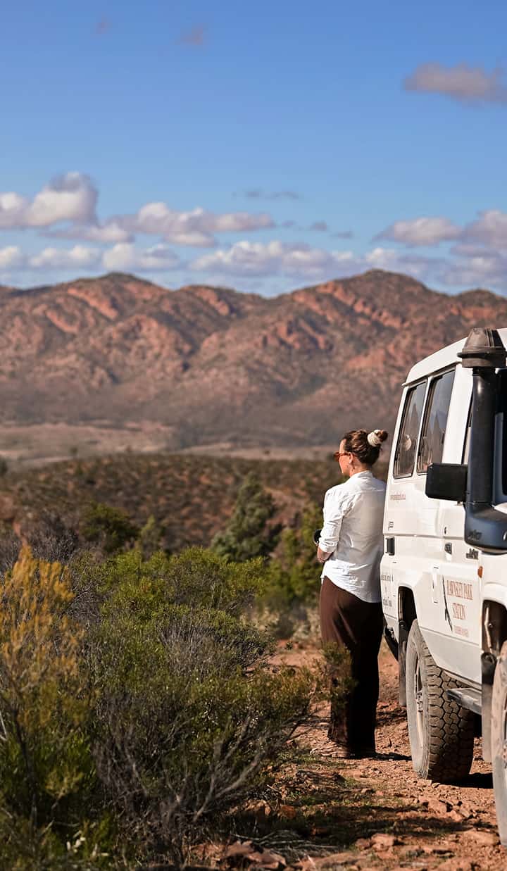 A woman stands beside an off‑road vehicle on a dirt track, looking out toward a wide landscape 