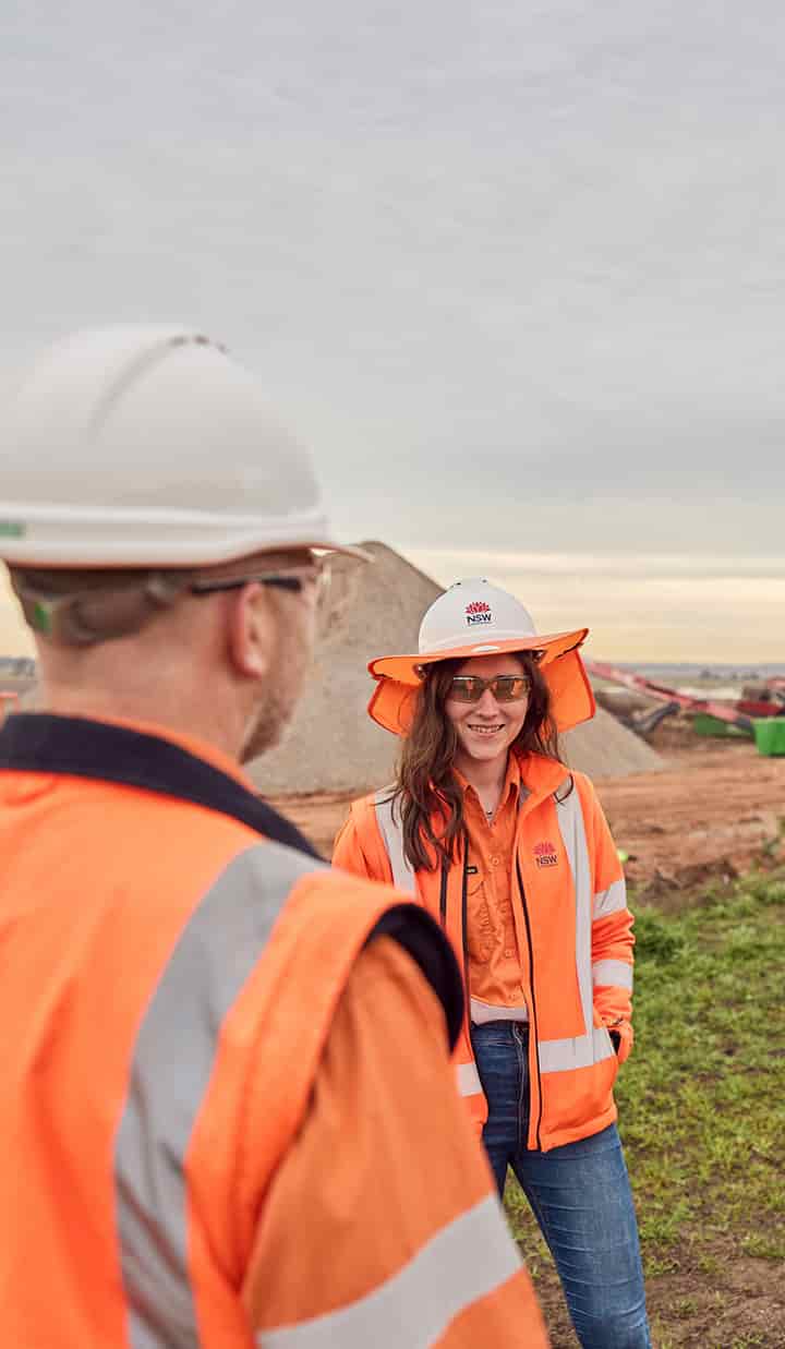 NSW workers in orange vests wearing PPE and standing in a construction site with a pile of sand in the background.