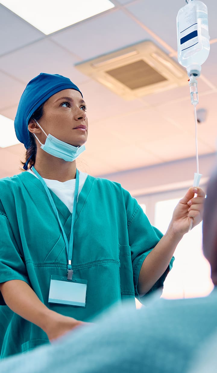 A medical professional wearing surgical scrubs, a cap, and a face mask is standing indoors, adjusting an IV drip bag.
