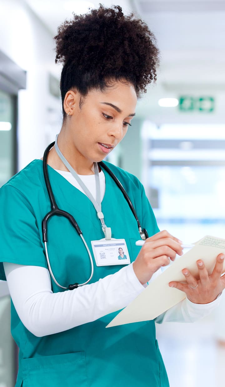 A healthcare professional wearing teal scrubs and a stethoscope around the neck is standing in a bright hallway, holding and writing on a clipboard