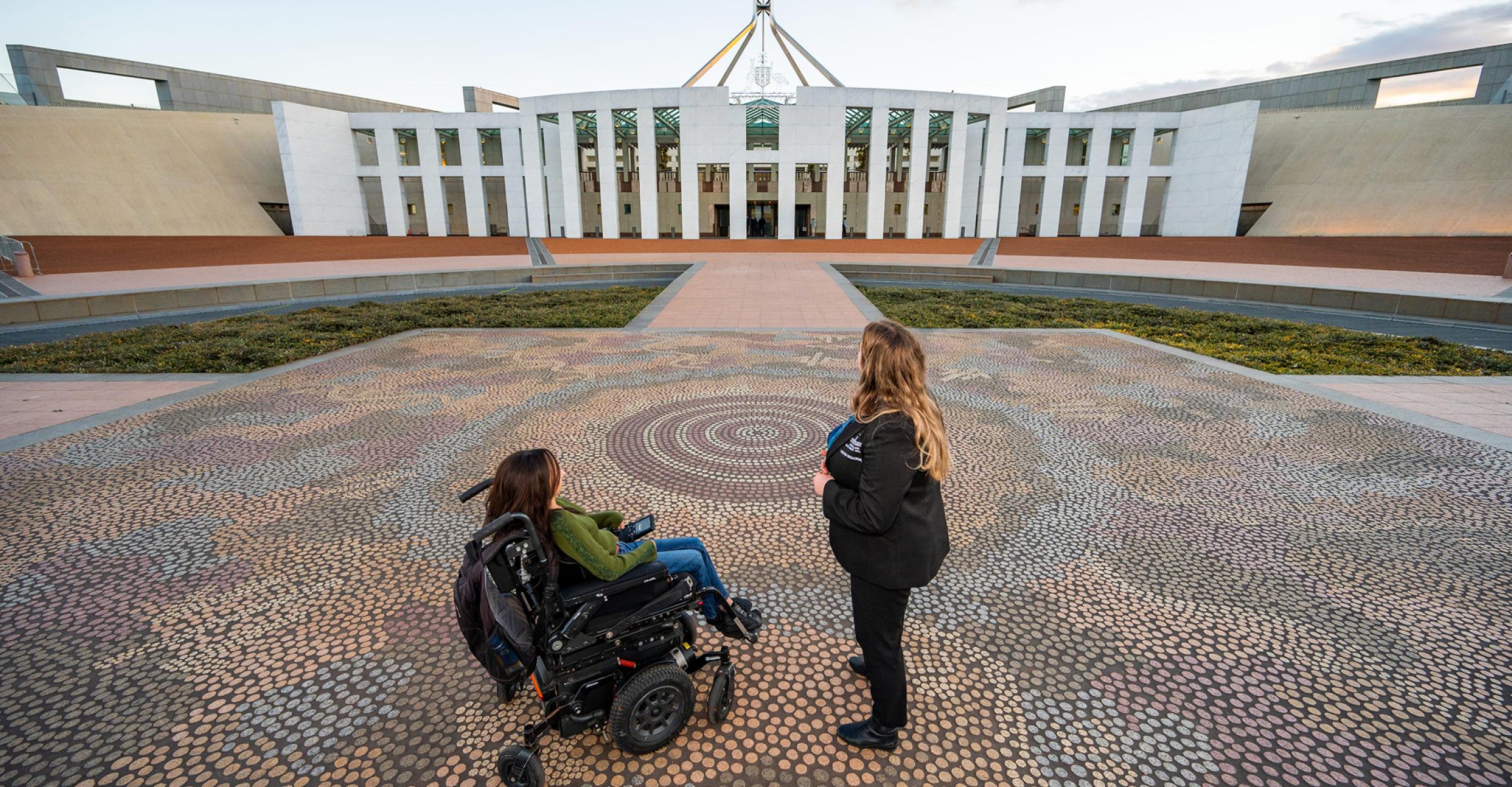 Person in a wheelchair and companion on Indigenous mosaic at Parliament House forecourt, Canberra
