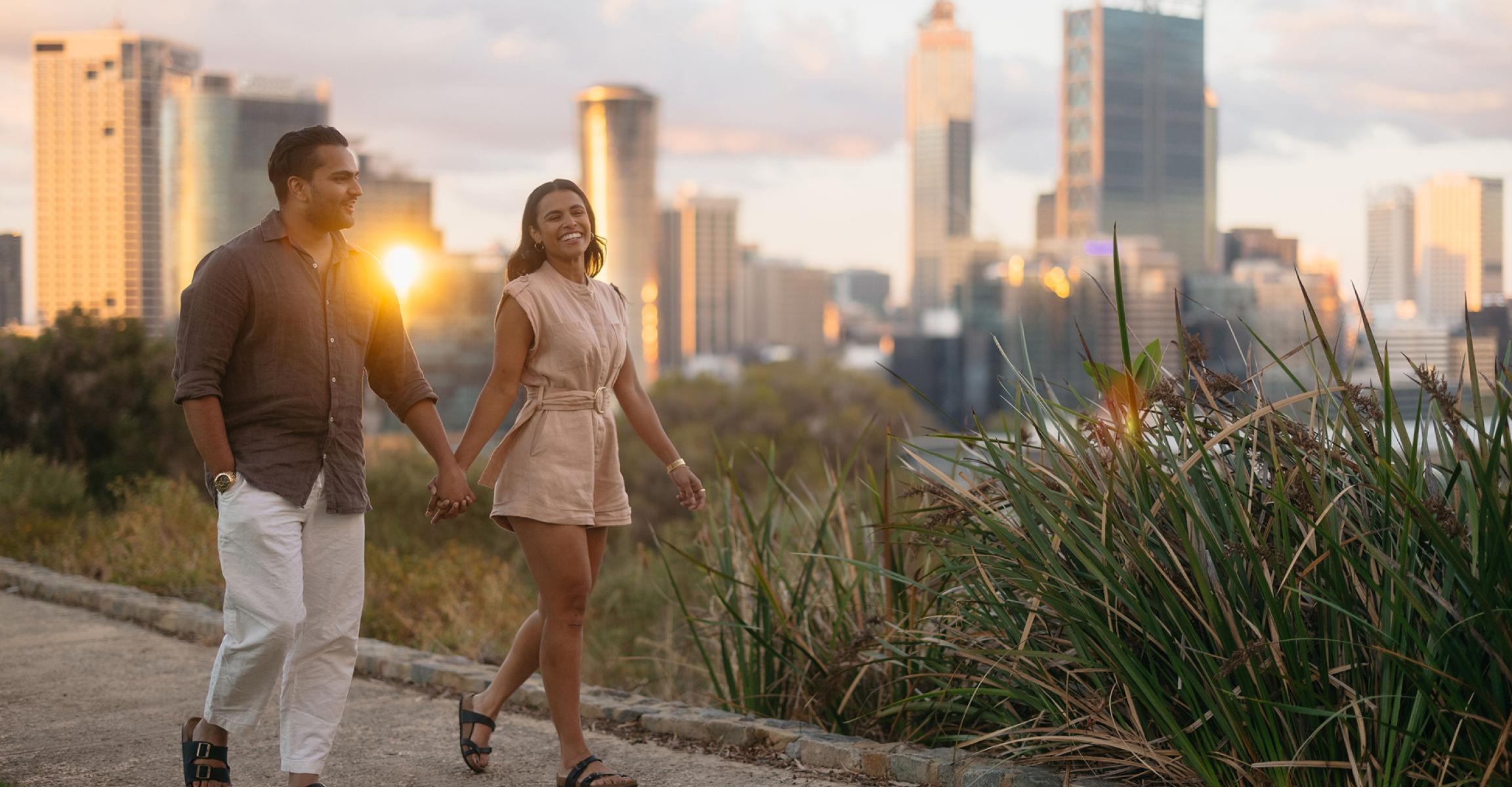 Couple holding hands strolling along a scenic path in Kings Park, Perth, with sunset city skyline in background.