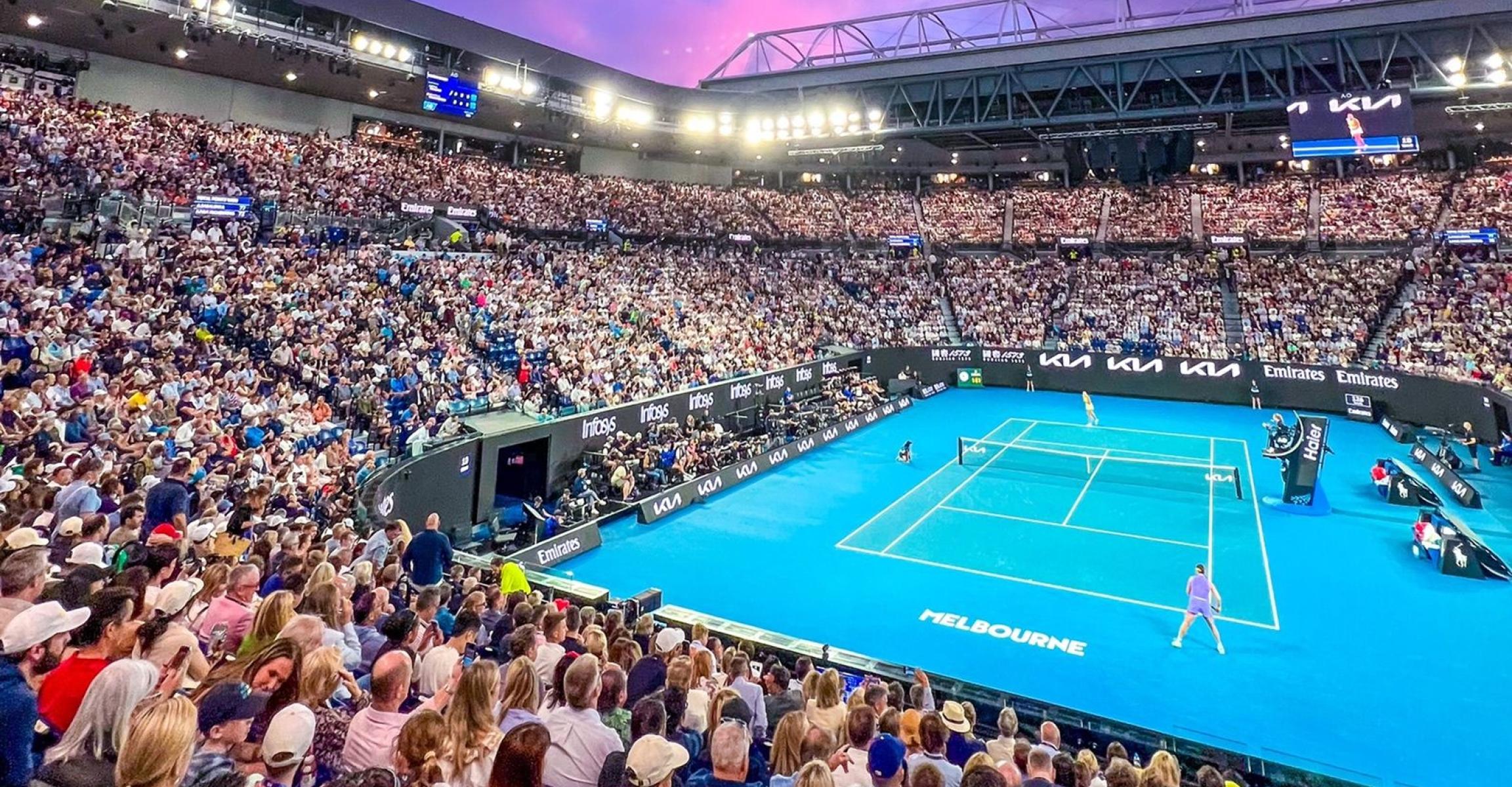 Crowd watching singles match at Australian Open, Melbourne Park, under floodlights with pink and purple sky.