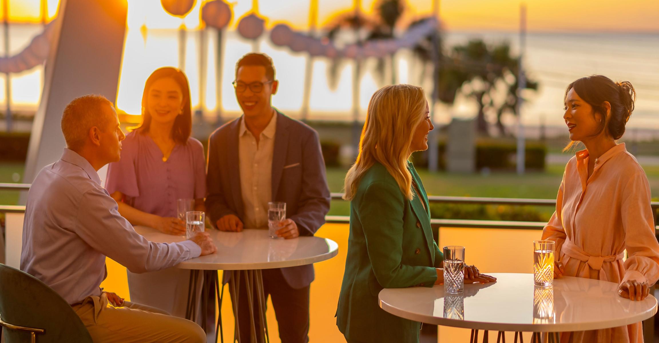Groups relaxing with drinks on the deck at Mindil Beach Casino Resort, Darwin, sunset over calm water and palm trees.