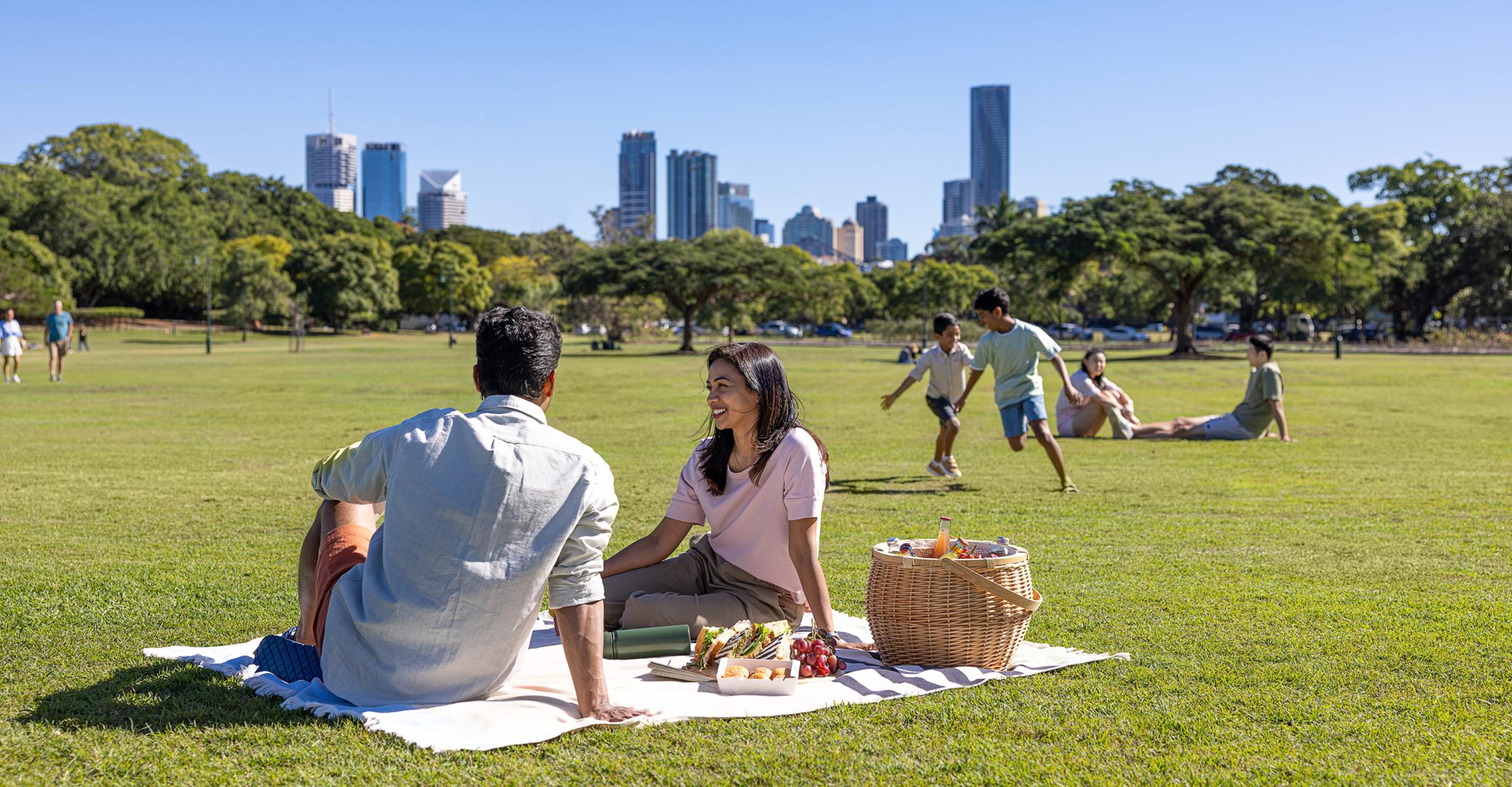 Family picnicking and relaxing in New Farm Park, Brisbane, with city skyline in the background.